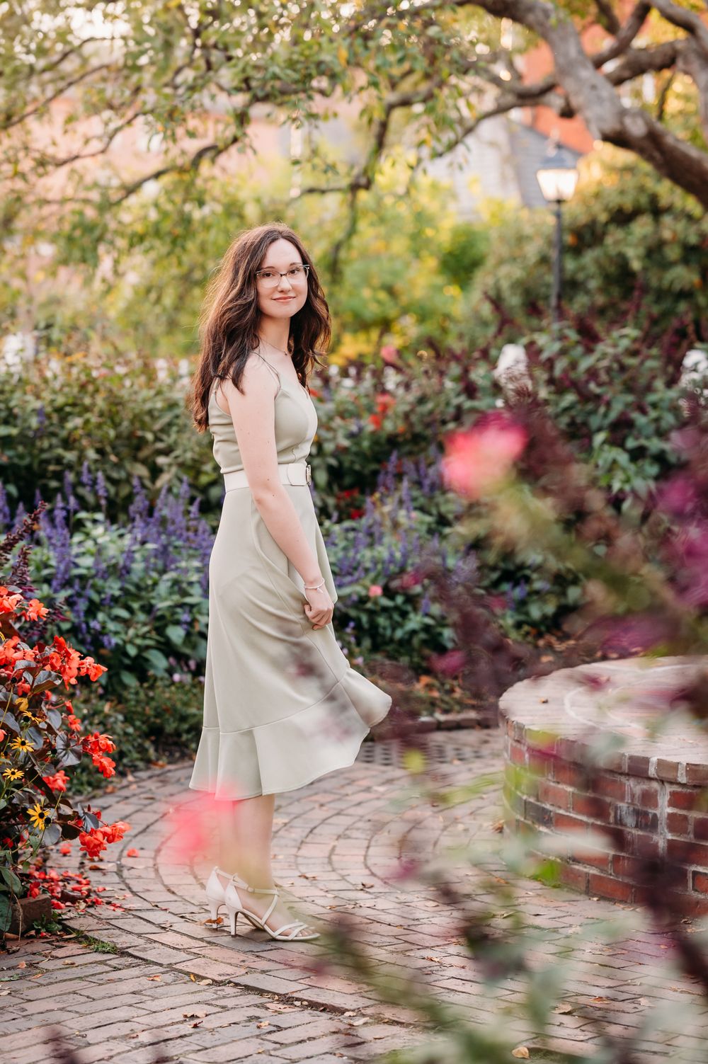 A green dress stands out against a colorful garden backdrop with brick pathways and blooming flowers in natural light.