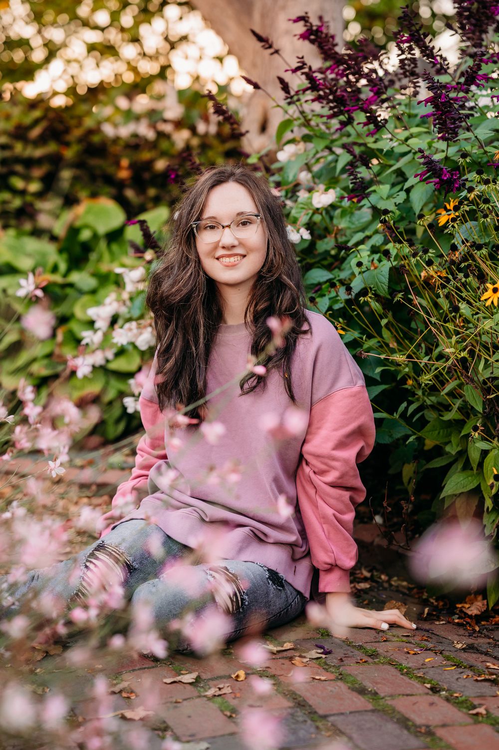 Person wearing pink sweater sits on brick path surrounded by vibrant garden plants and bokeh lighting at sunset.