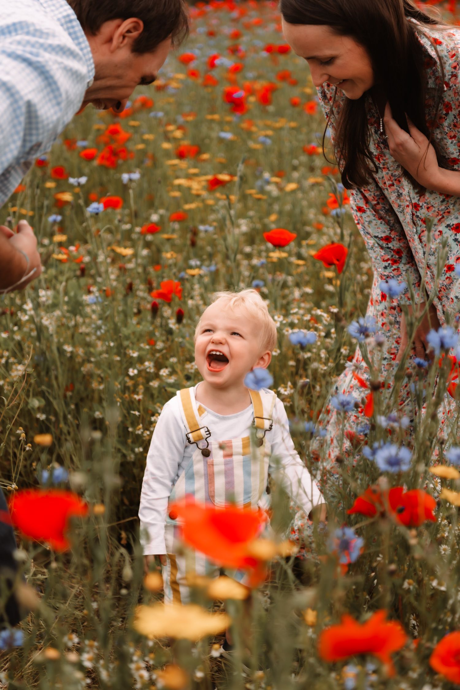 A joyful young child in checkered overalls laughs while standing in a vibrant field of red poppies and wildflowers.