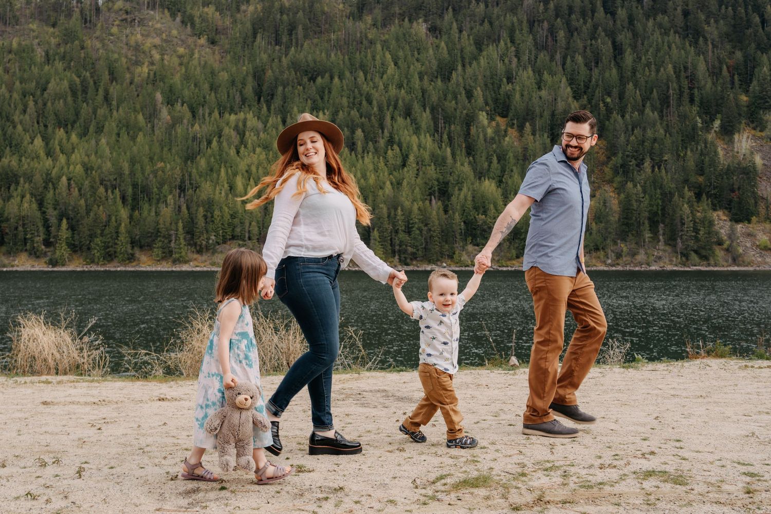 A family holds hands while walking along a scenic lakeside beach with mountains and forest in the background.