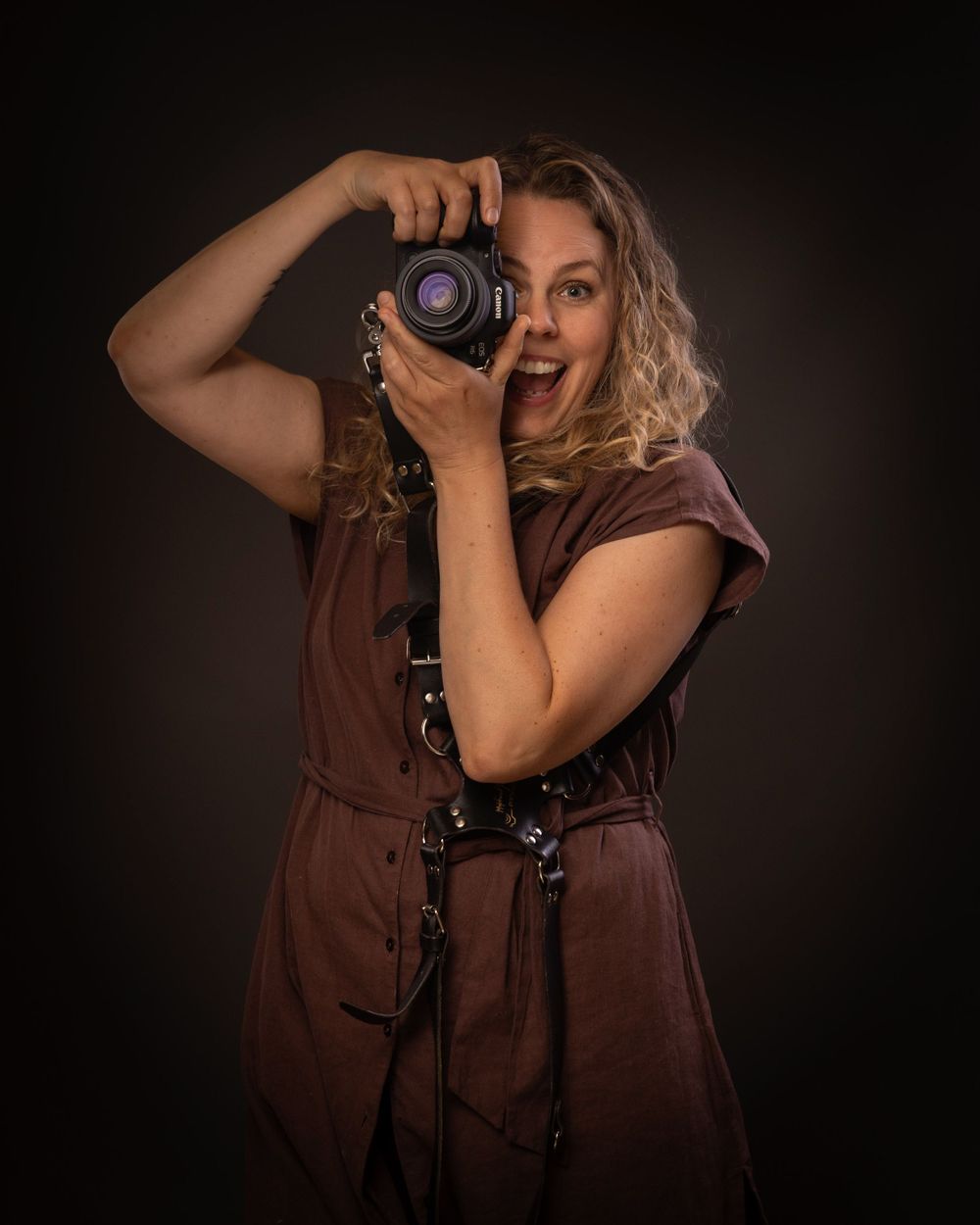 Professional photographer in brown dress holding camera with excited expression against dark background.