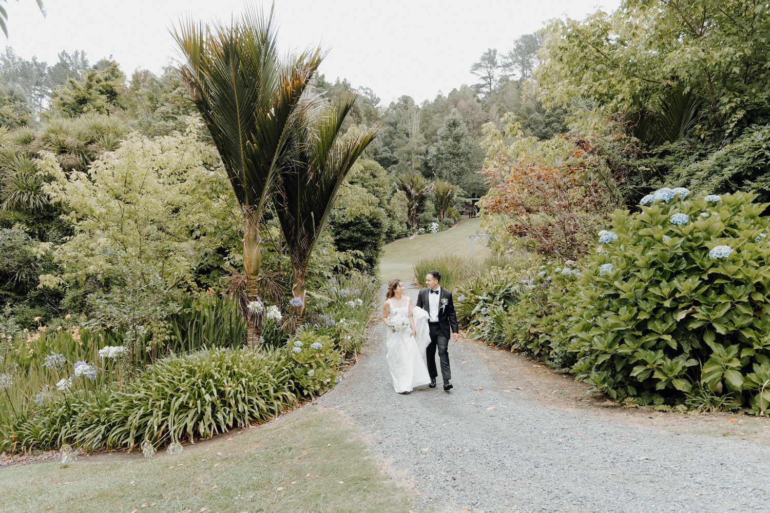 Bride and groom walk hand in hand down a gravel path surrounded by lush gardens captured by Apertured Media Auckland Wedding Photographer.