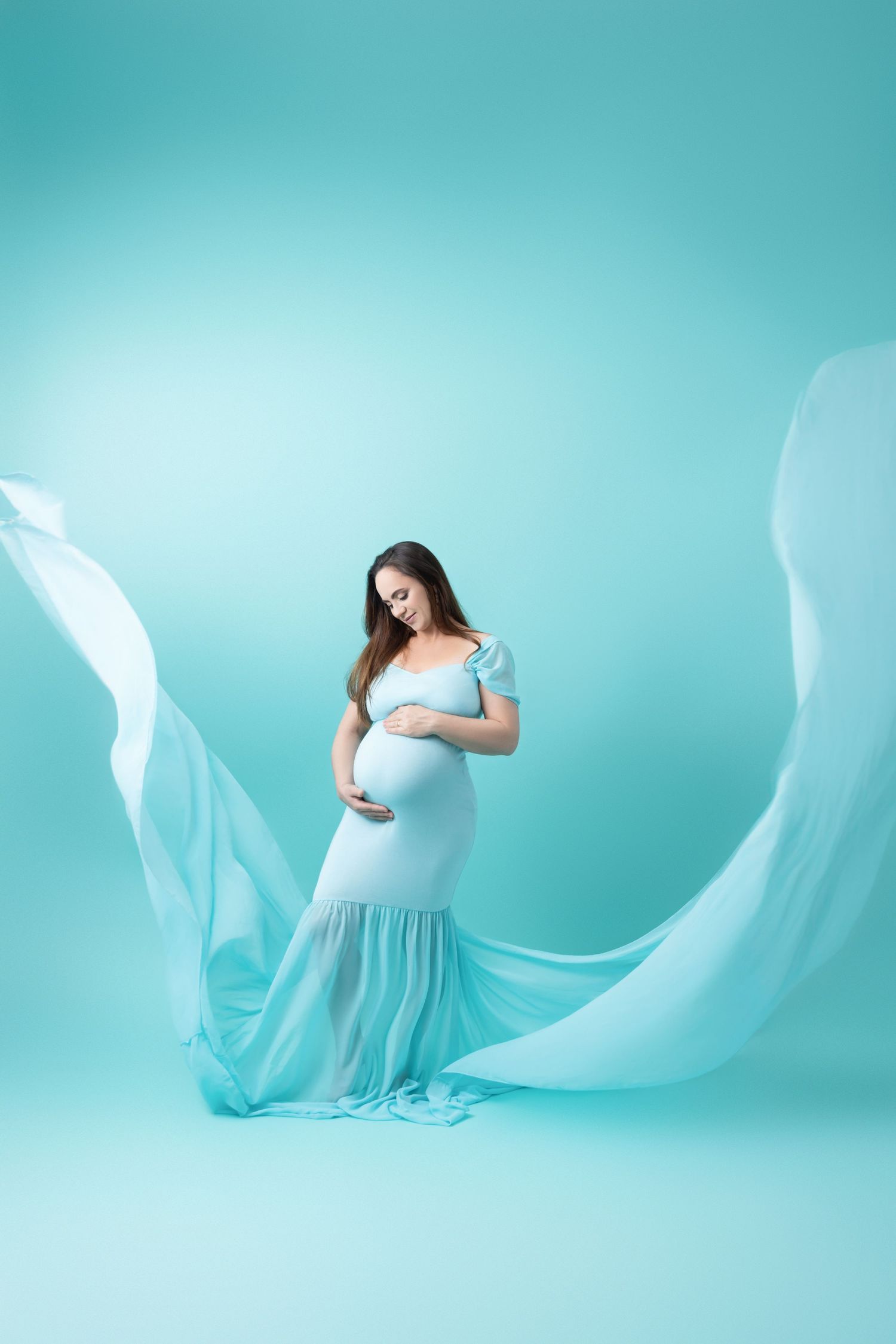 A serene maternity photo with flowing turquoise fabric against a teal background creating an ethereal effect.