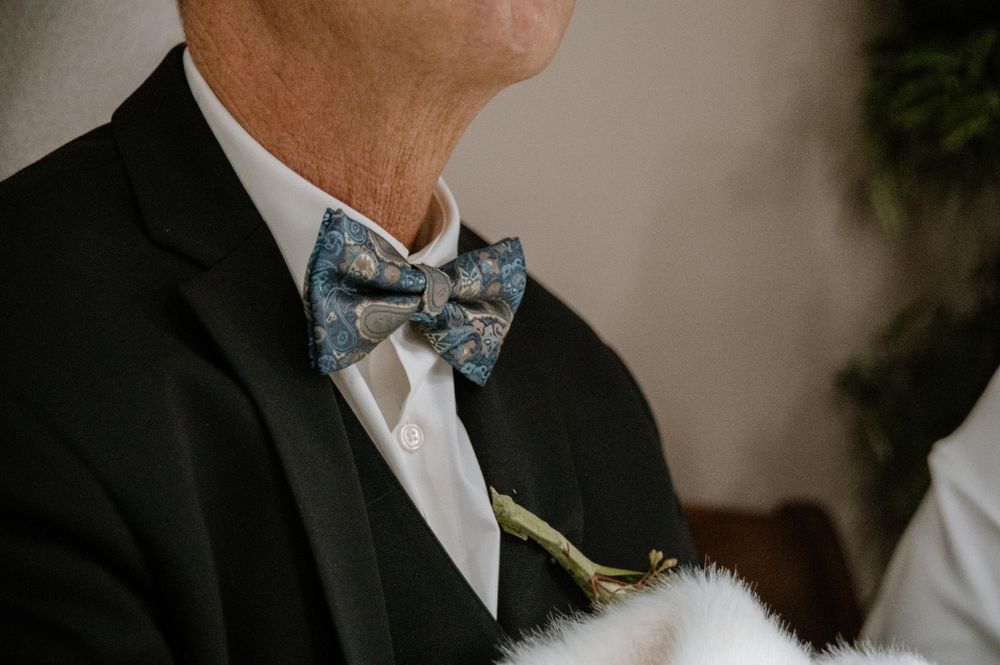 Close-up of a blue floral bow tie and black suit jacket with white boutonniere at a formal event.