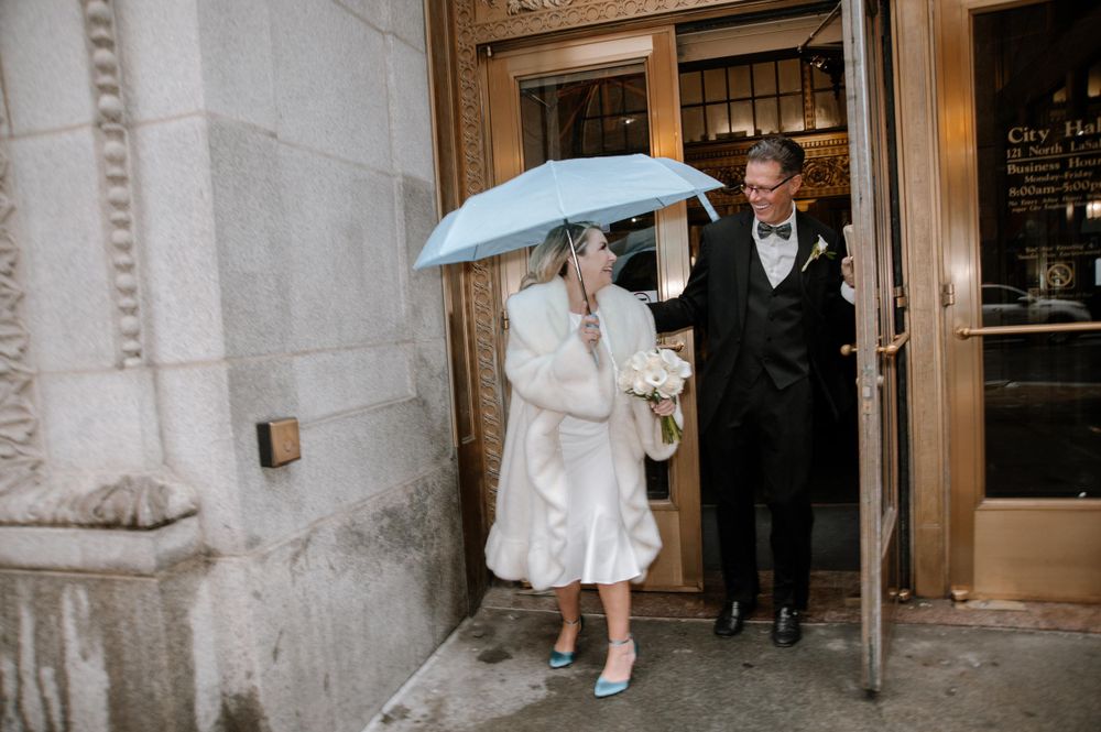 A couple shelters under a light blue umbrella outside an elegant building entrance on a rainy day.