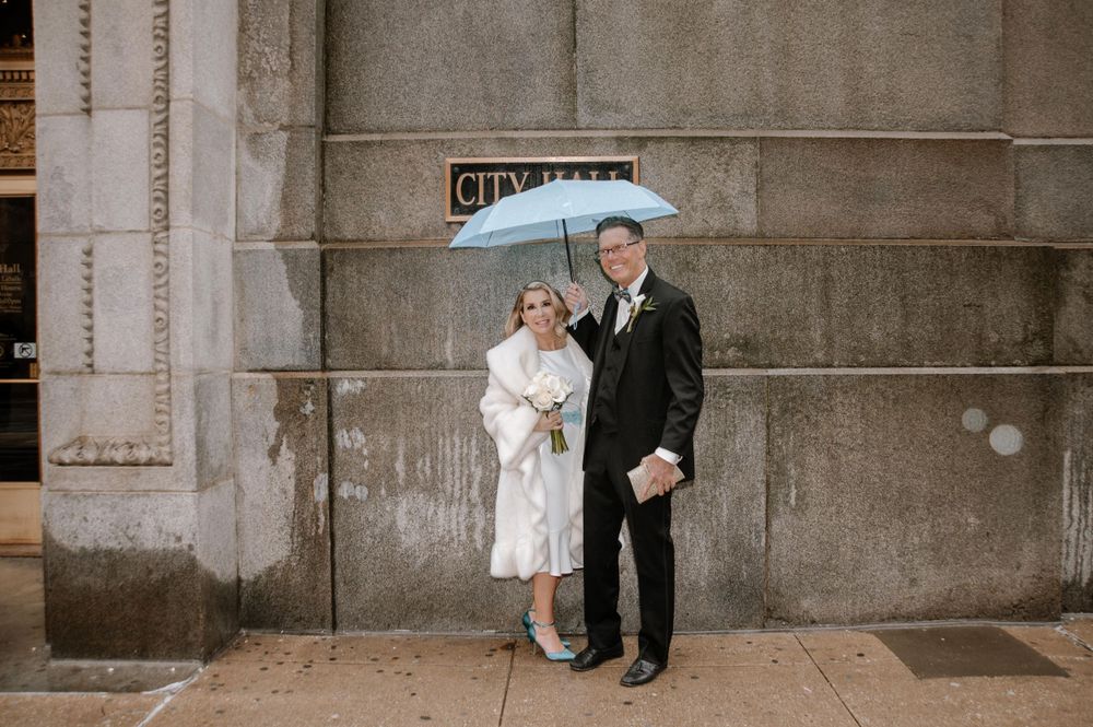 A couple shares a light blue umbrella while dressed in wedding attire against an urban concrete wall.