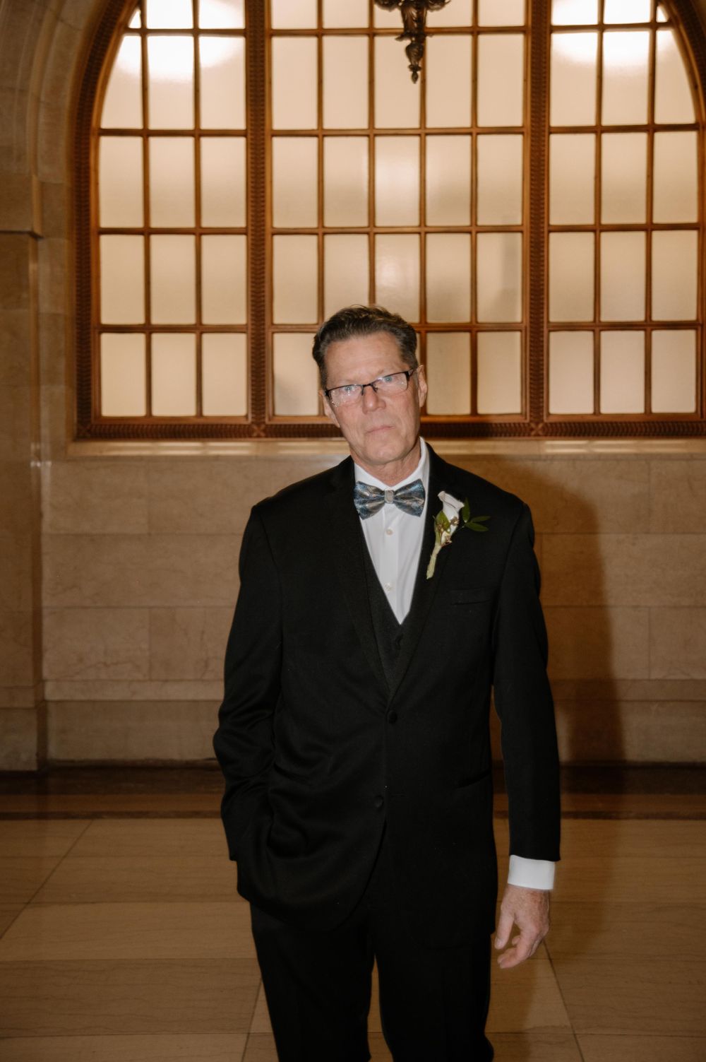 A formal event attendee in a black tuxedo and bow tie stands in front of a large windowed wall in warm lighting.