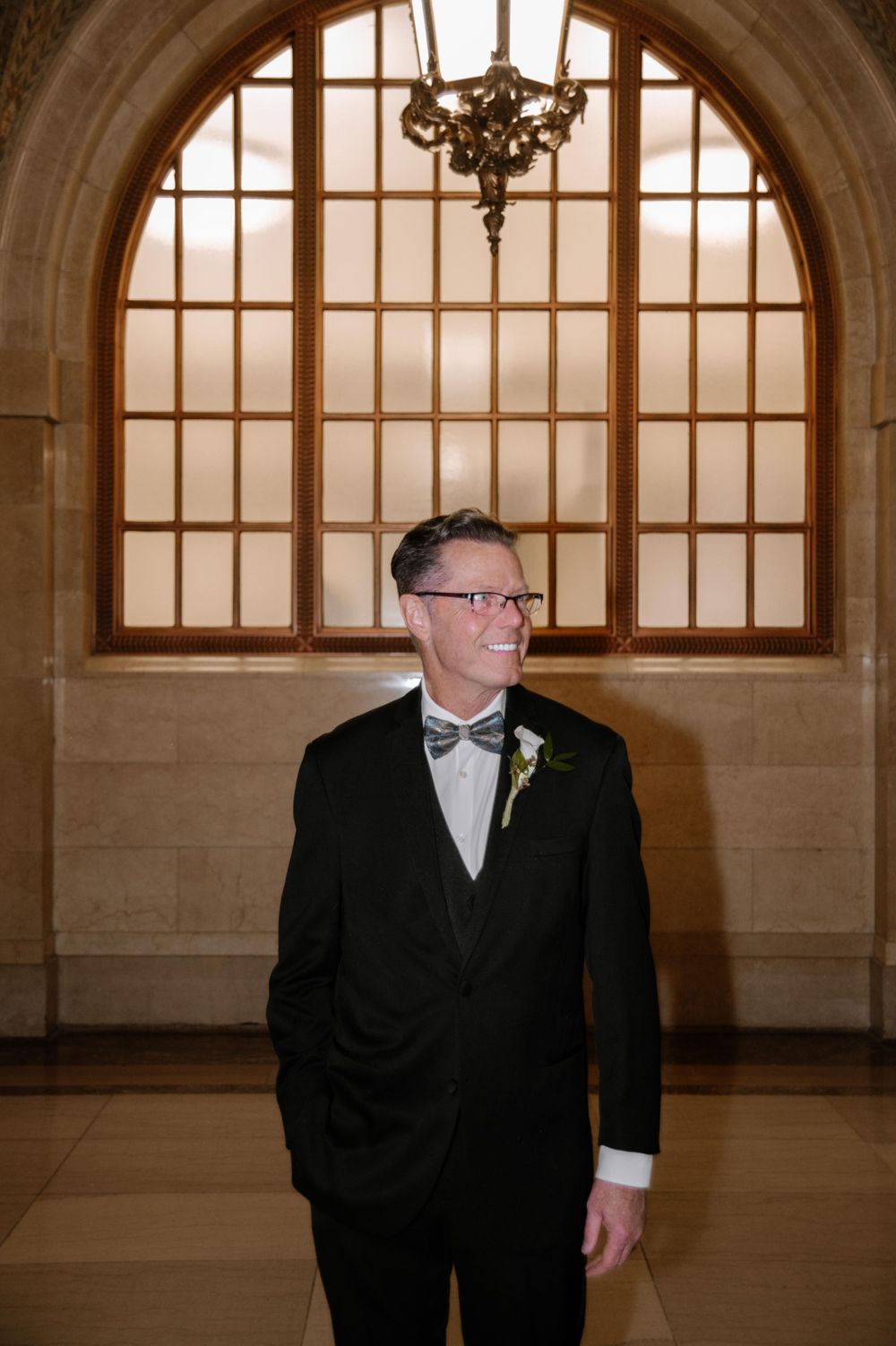 A groom in a black tuxedo and bowtie stands in front of an elegant arched window with warm backlighting.