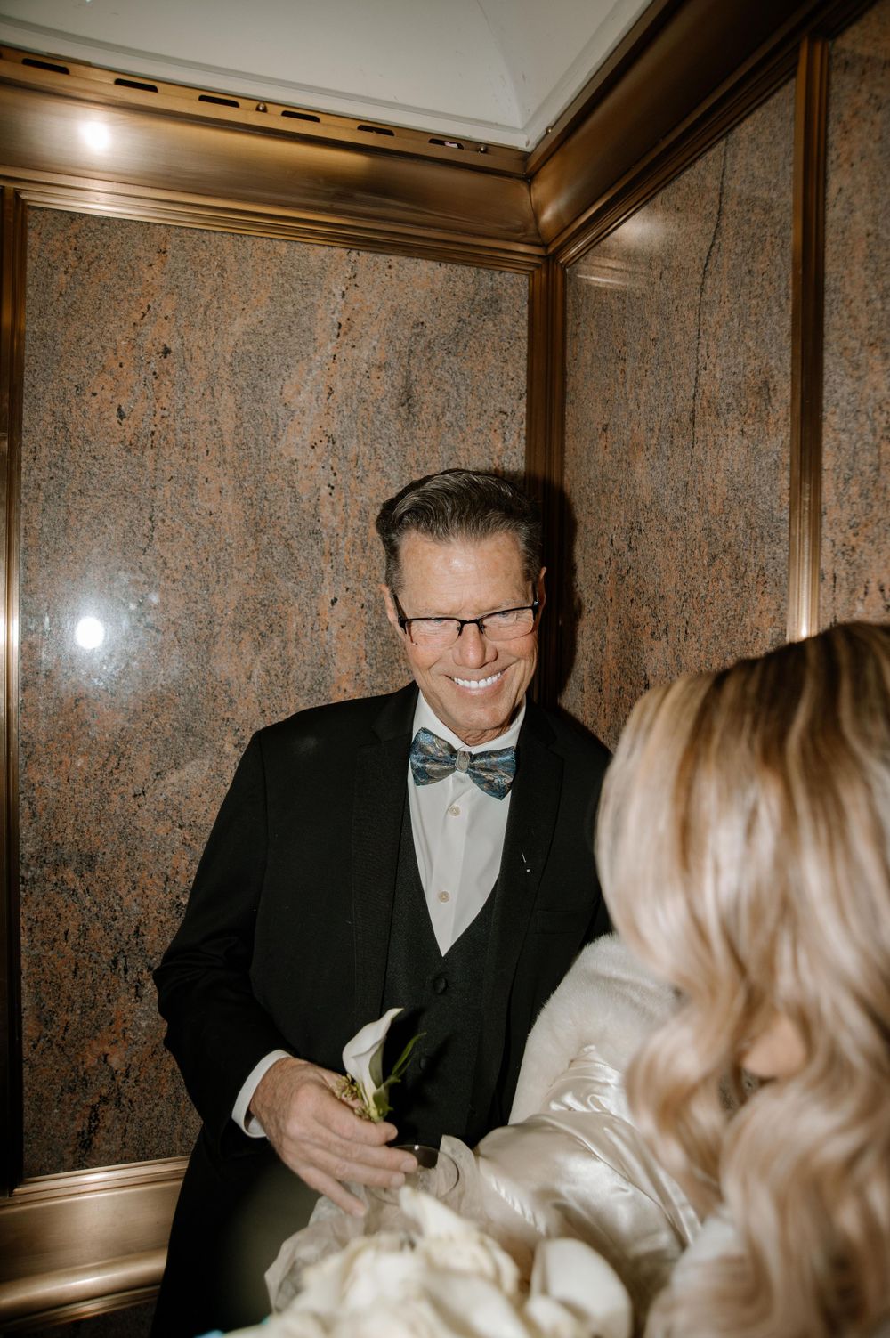 Wedding guest in formal attire with bowtie and glasses smiles warmly during an indoor evening celebration.
