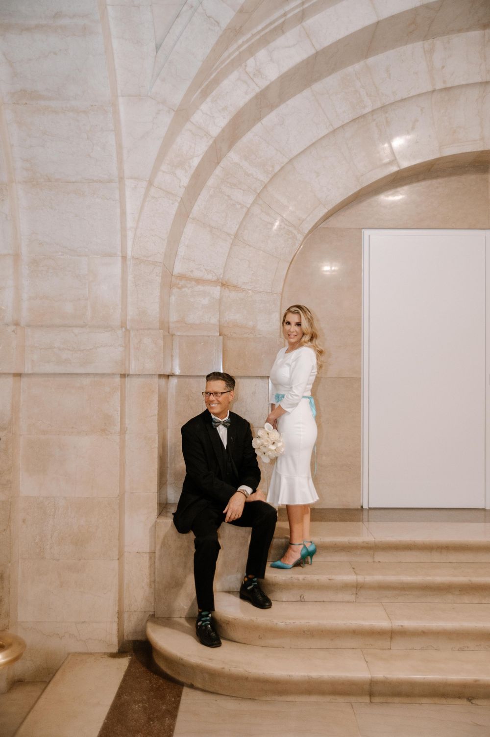 Couple in wedding attire poses elegantly on curved stone steps under arched architectural details.
