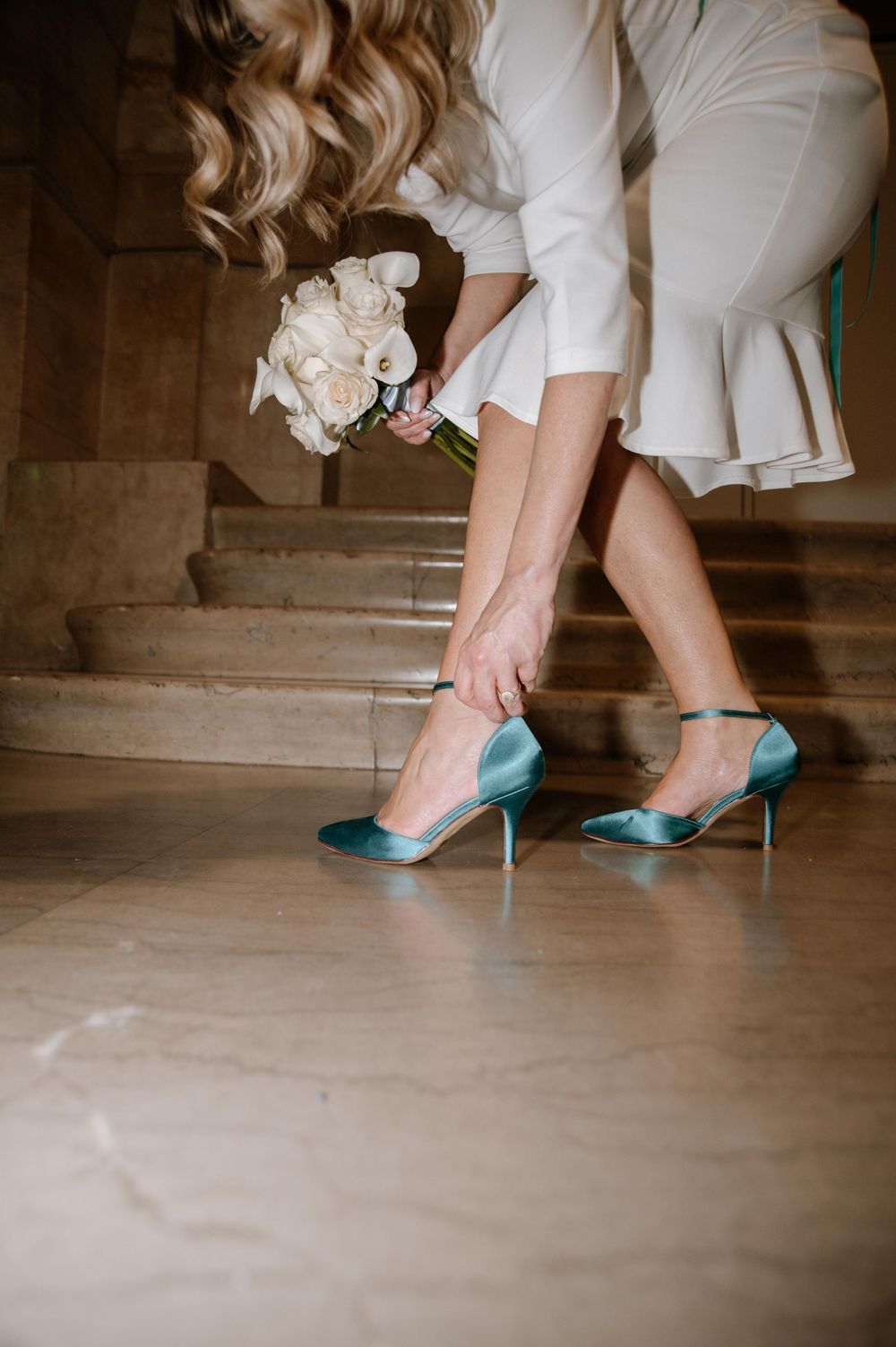 A bride in white dress adjusts her teal high heels on stone steps while holding a bouquet.