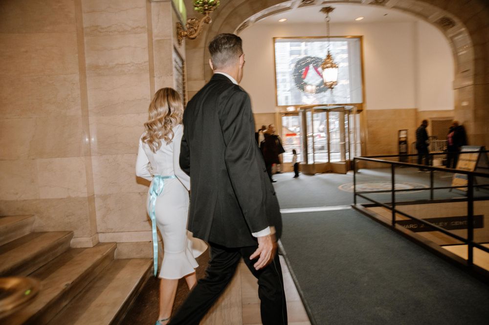 Well-dressed couple walking down carpeted hallway in ornate building interior with arched entryway.