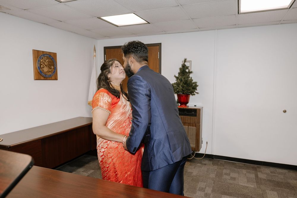 A couple shares a tender moment in an orange traditional dress and blue suit at a small indoor ceremony.