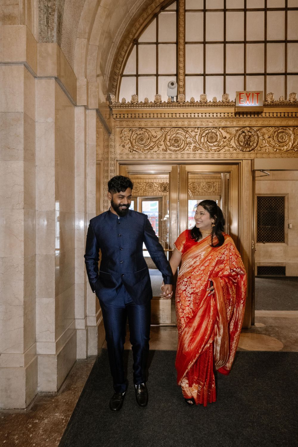 A couple in traditional South Asian attire, with navy suit and red sari, walking together in an ornate hallway.