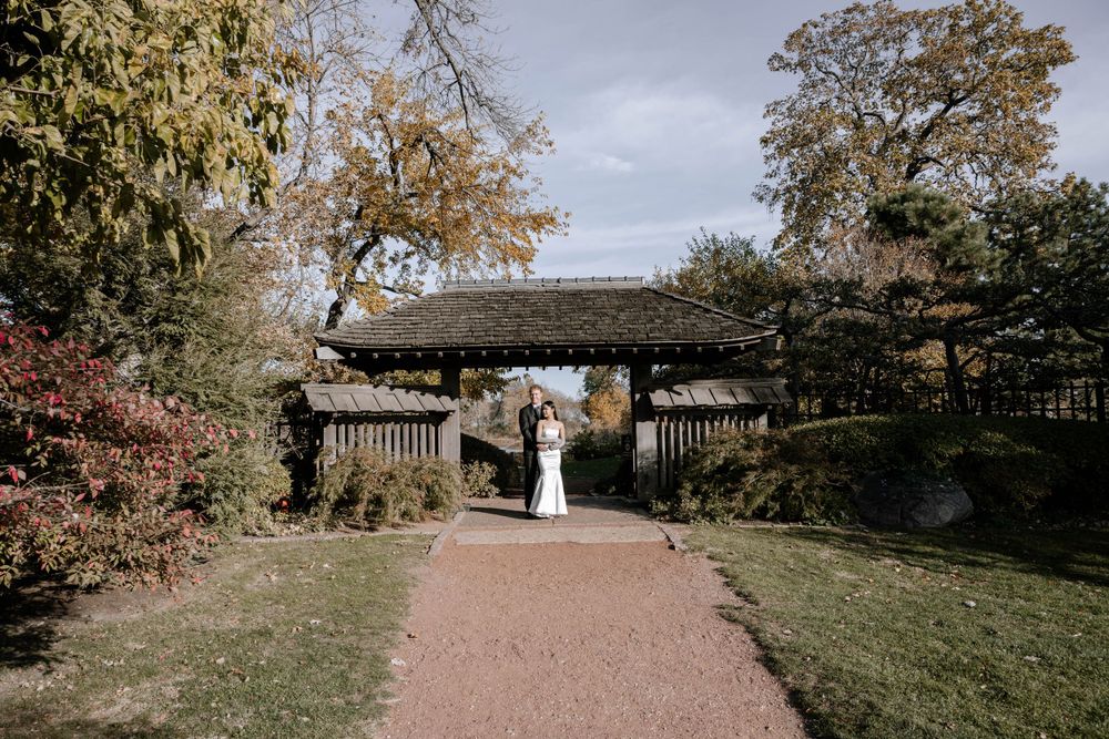 A couple stands under a rustic wooden pergola on a brick path surrounded by autumn trees and grass in a garden setting.