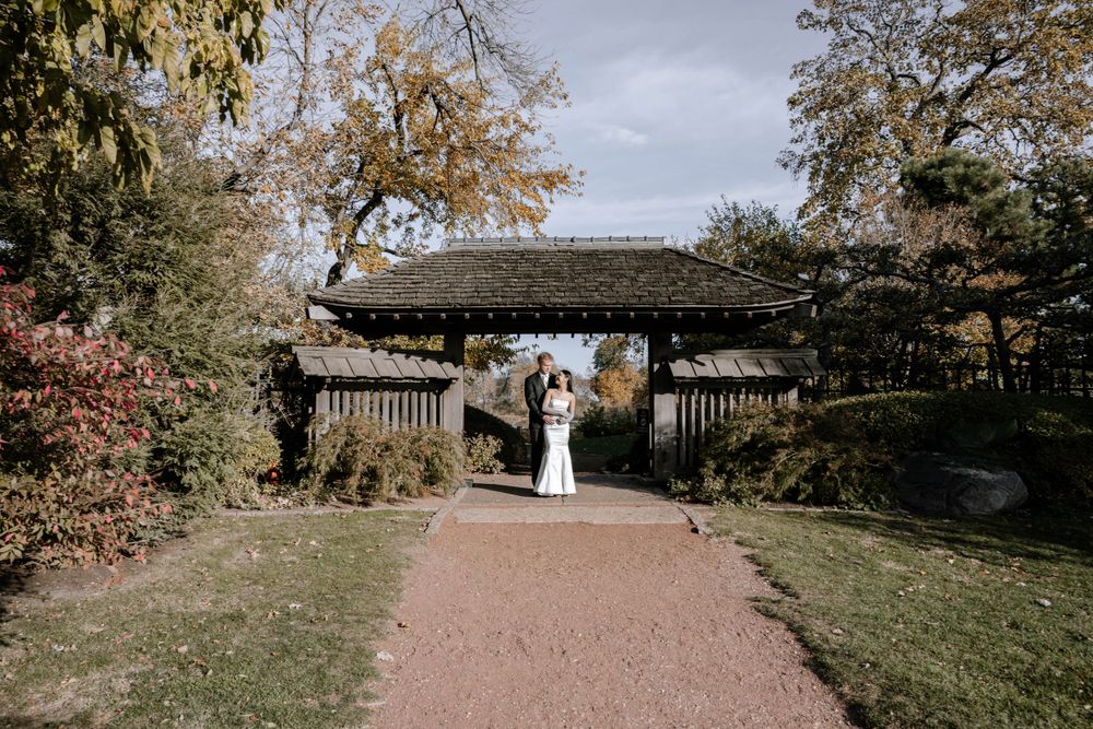 A couple stands under a rustic wooden pergola on a gravel path surrounded by autumn trees and foliage.