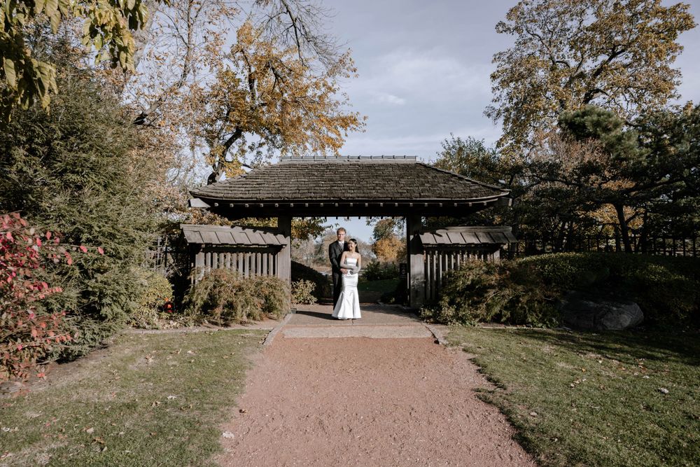 A wedding moment captured beneath a wooden pergola structure along a brick pathway in a fall garden landscape.