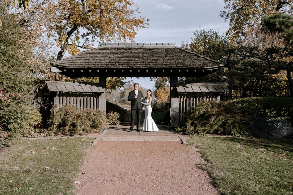 A wedding couple stands beneath a traditional Japanese wooden arch gate on a garden path during autumn.