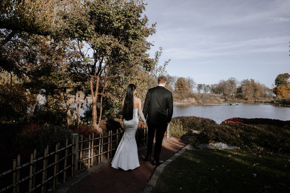 A couple stands together on a lakeside path at sunset with autumn trees and water in the background.