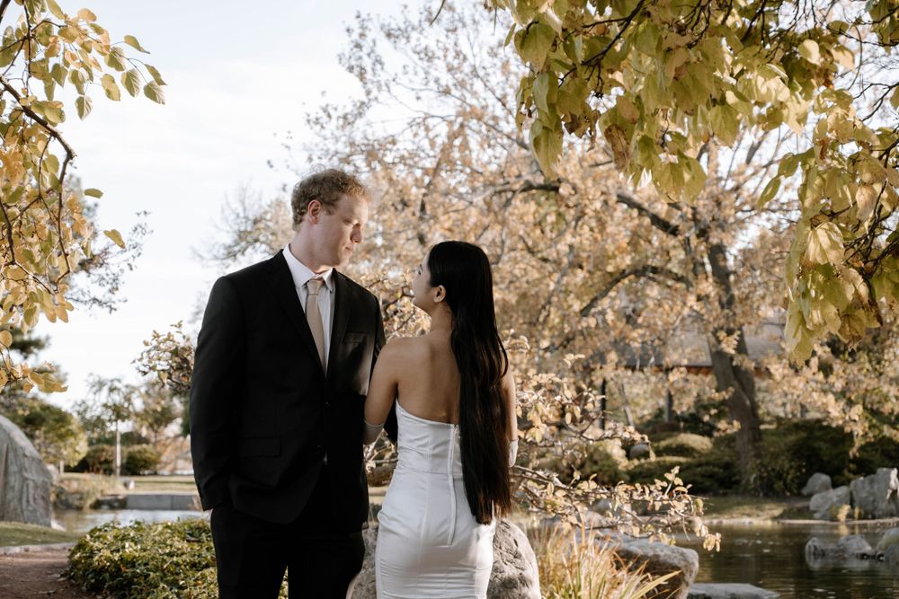 A couple in formal wedding attire share a romantic moment in a garden setting with autumn foliage.