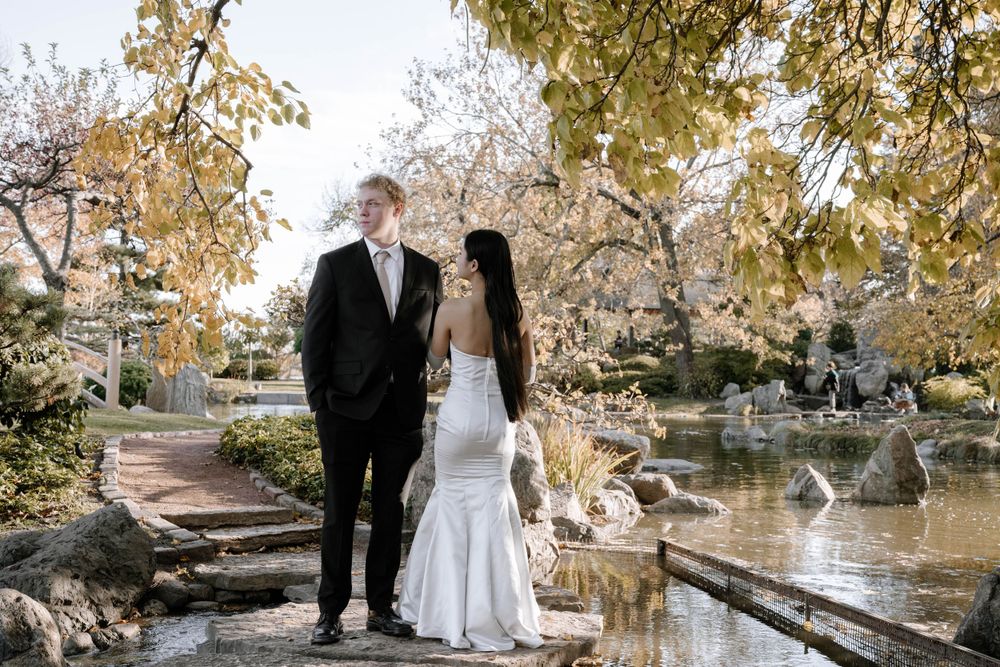 A couple in wedding attire stands elegantly by a scenic pond surrounded by autumn trees and rocks.