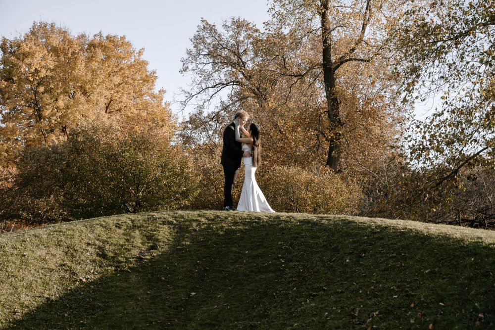 A couple in wedding attire stands on a grassy hilltop surrounded by autumn trees with golden foliage.