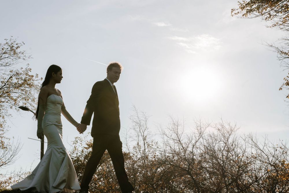 A romantic silhouette of a bride and groom walking hand in hand against a bright sky at sunset.