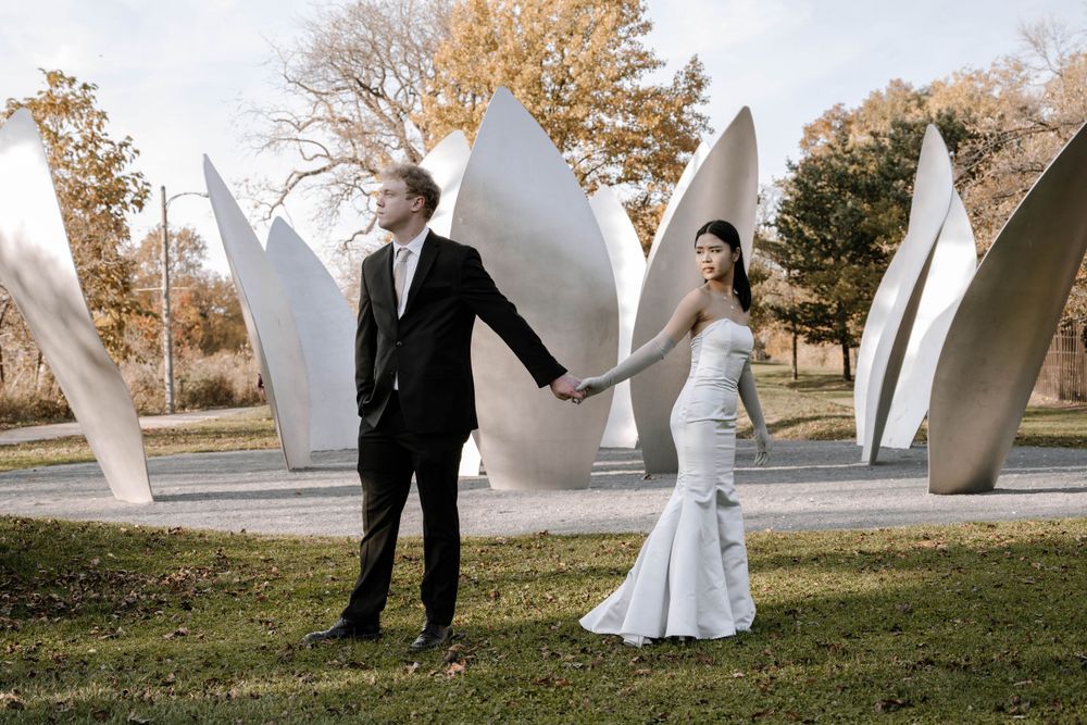 A couple in wedding attire posing in front of large white sculptural petals in an outdoor garden setting.