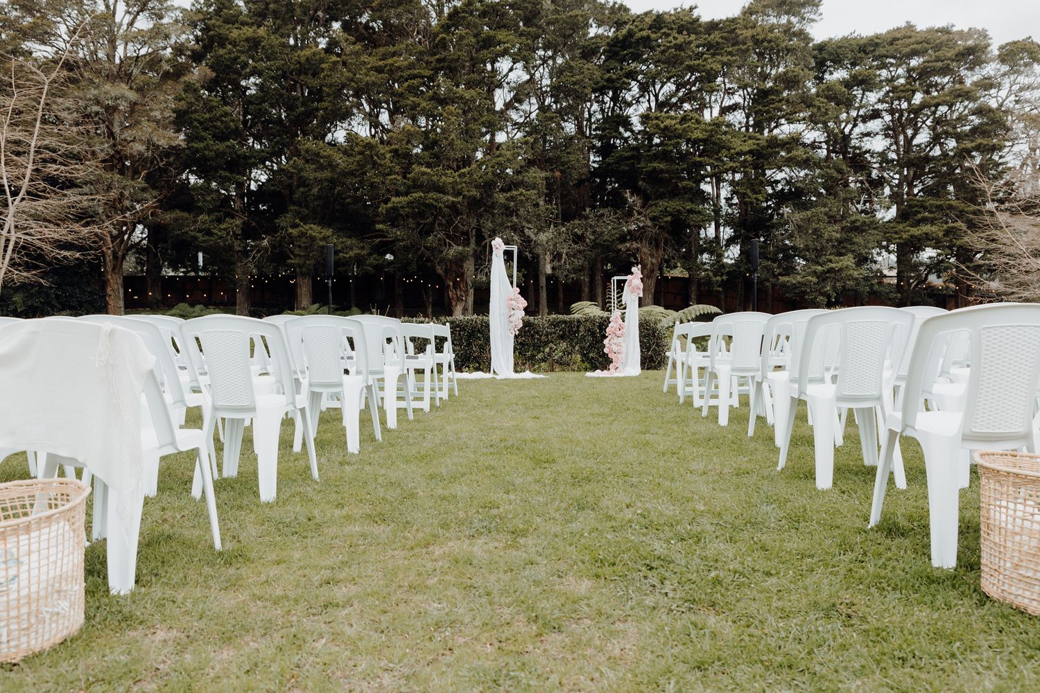 Garden wedding ceremony setup with white chairs and wicker baskets lining the aisle.