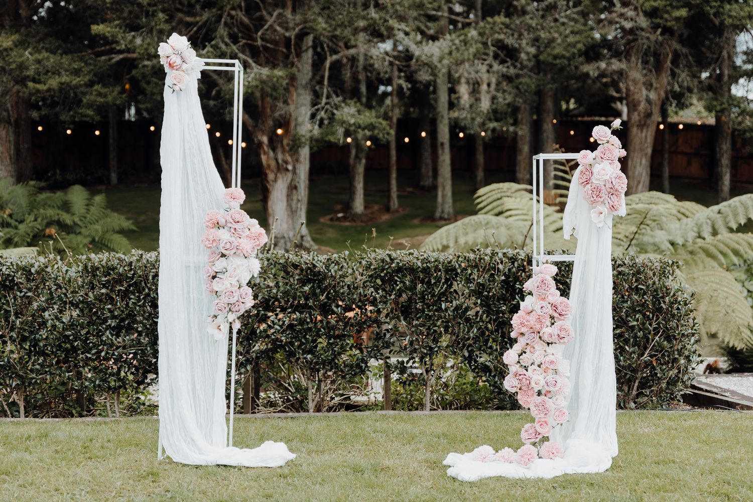 White wedding arch decorated with pink floral arrangements in an outdoor garden setting.