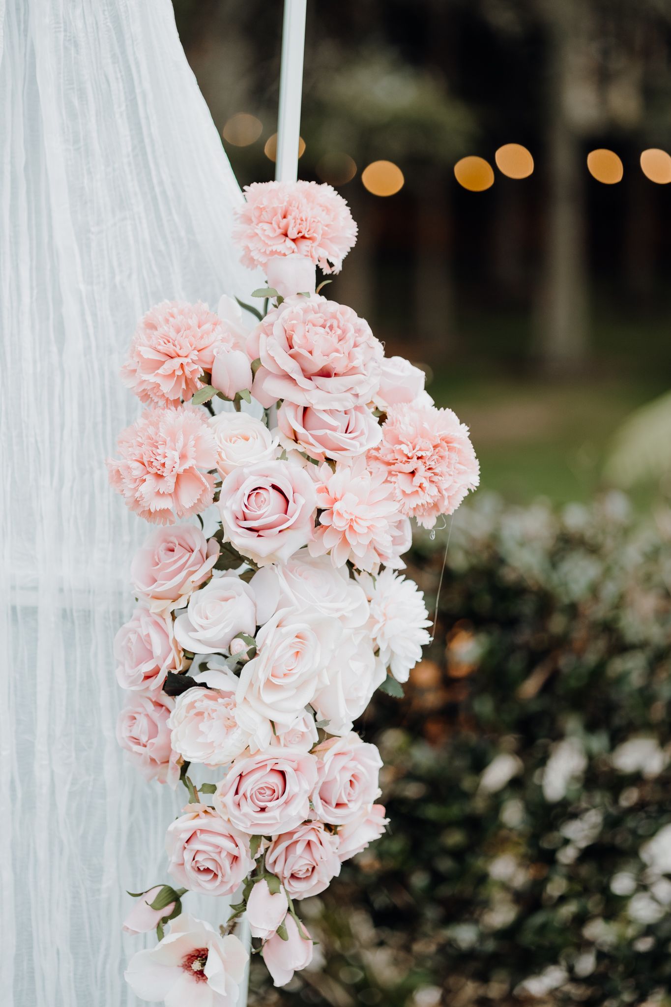 Close-up of delicate pink roses and carnations arranged on white fabric with bokeh lights in background.