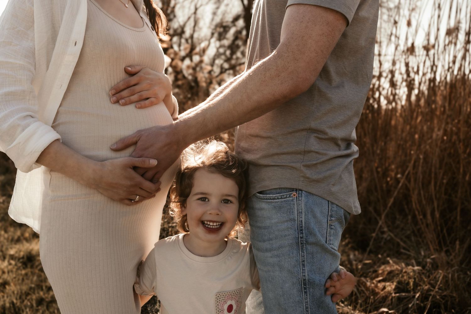 A joyful child stands between expecting parents in a golden sunlit field at sunset.