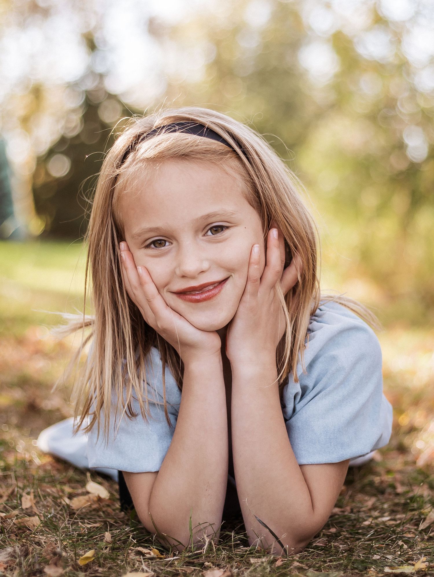 Portrait photo outdoors with natural bokeh background showing someone lying on ground with hands on cheeks in casual attire.