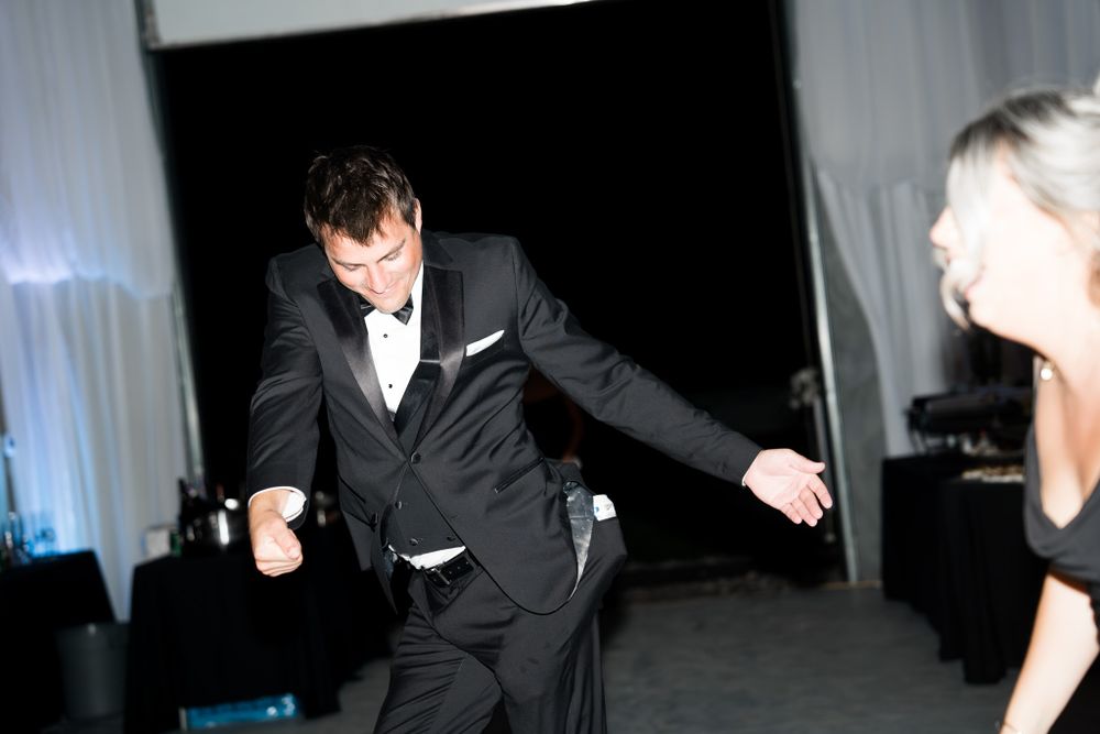 Someone wearing a black tuxedo dances energetically at an evening event with white curtains in the background.