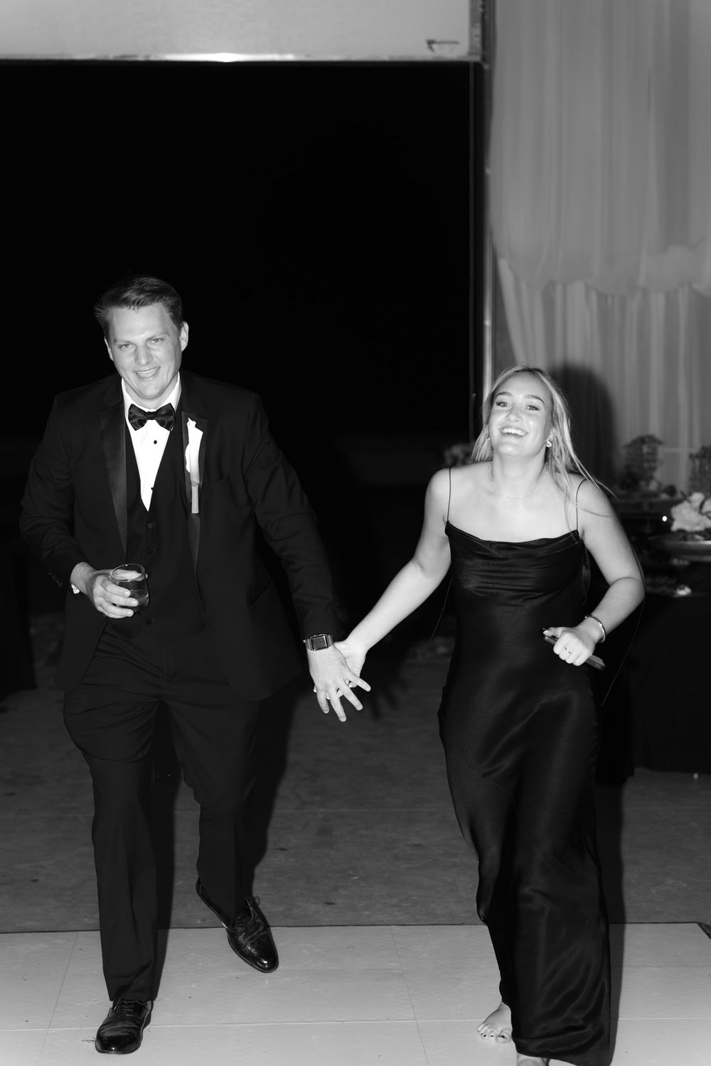 A couple in formal black attire walk together and laugh during an elegant black-tie event.