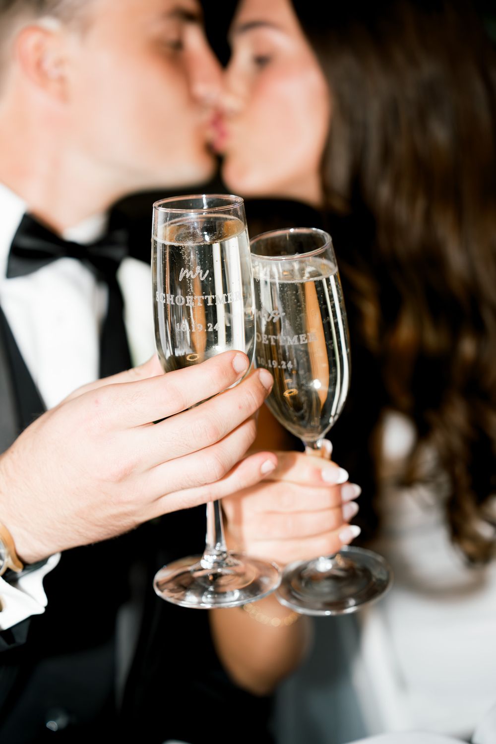 Two champagne flutes clink together in celebration as a couple shares an intimate moment in formal attire.