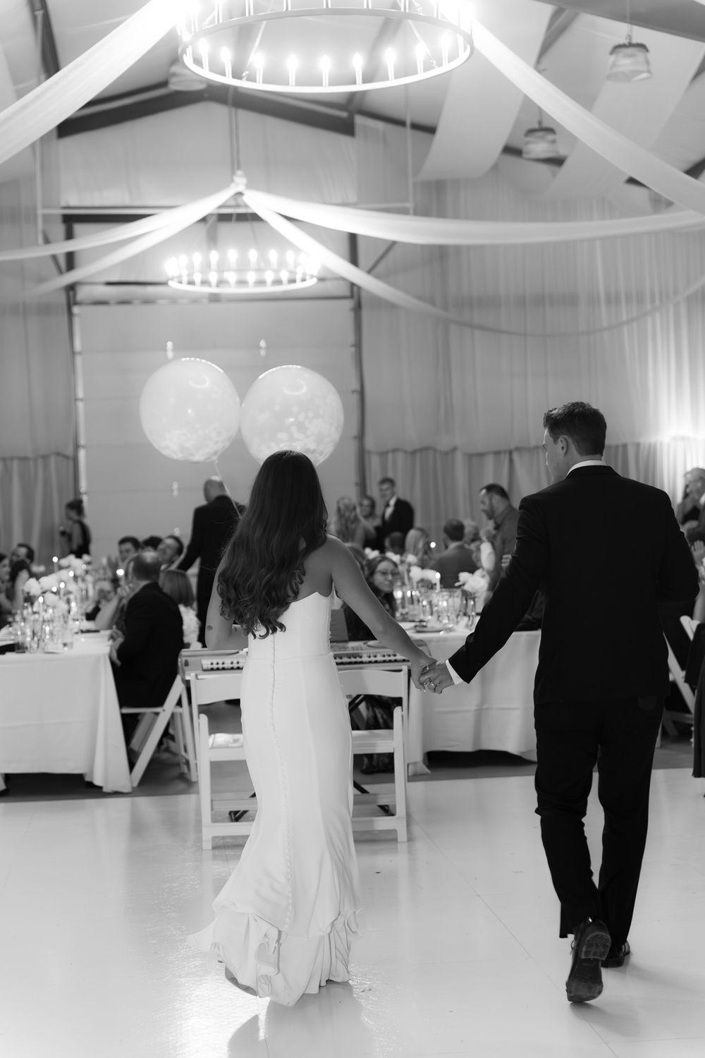 A couple holding hands while entering an elegant wedding reception venue decorated with white drapes and chandeliers.
