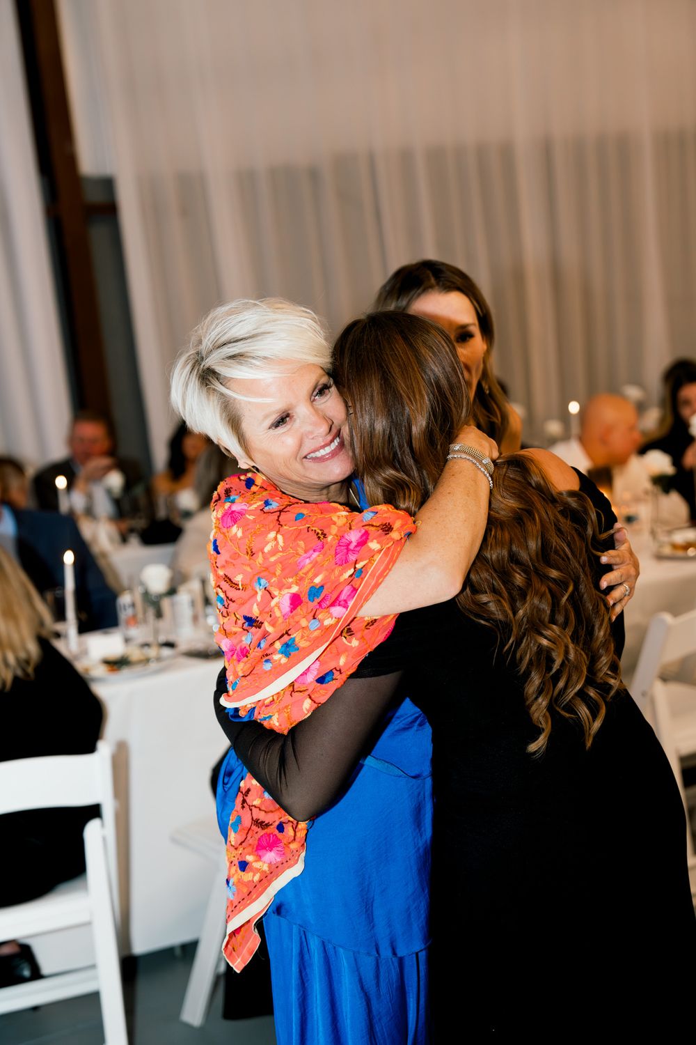 Warm embrace between wedding guests wearing vibrant orange and blue dresses at a reception celebration.