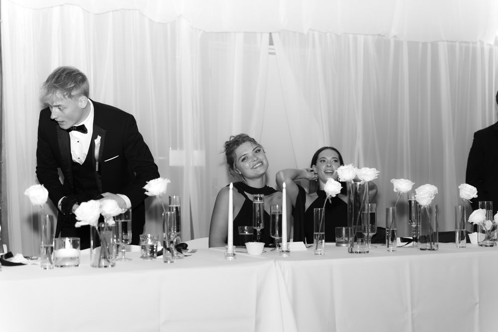 Black and white photo of formal banquet with people seated at table with drinks and white floral centerpieces.