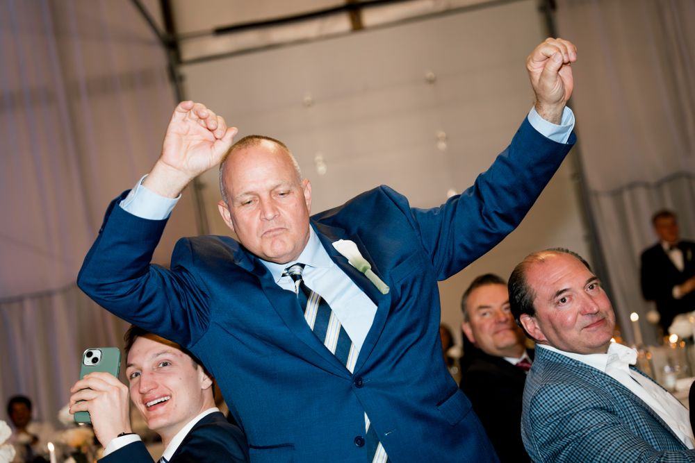Enthusiastic guest in blue suit raises arms in celebration during a wedding reception.