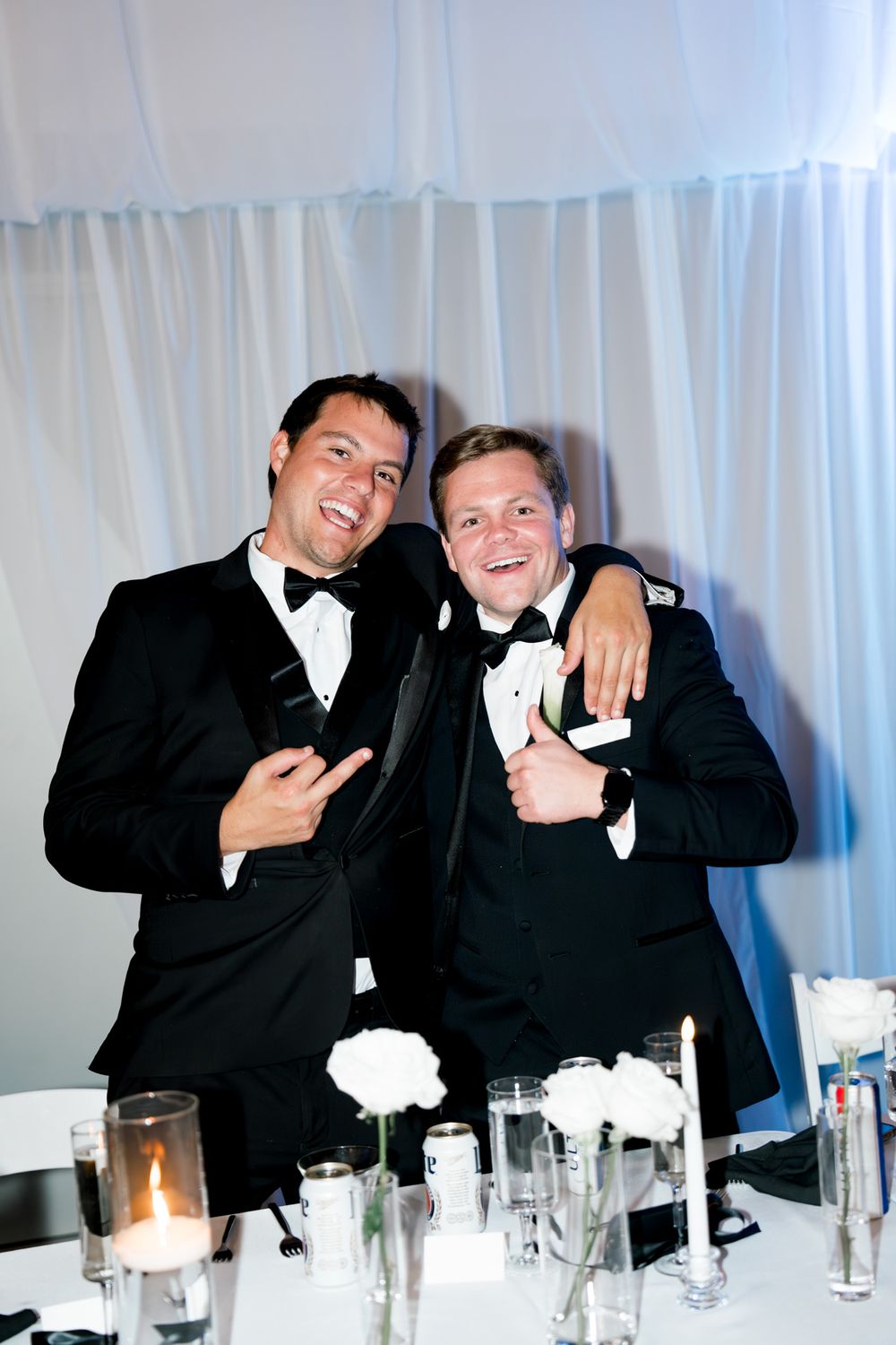 Two friends in tuxedos pose together at an elegant reception with table settings and white decor.