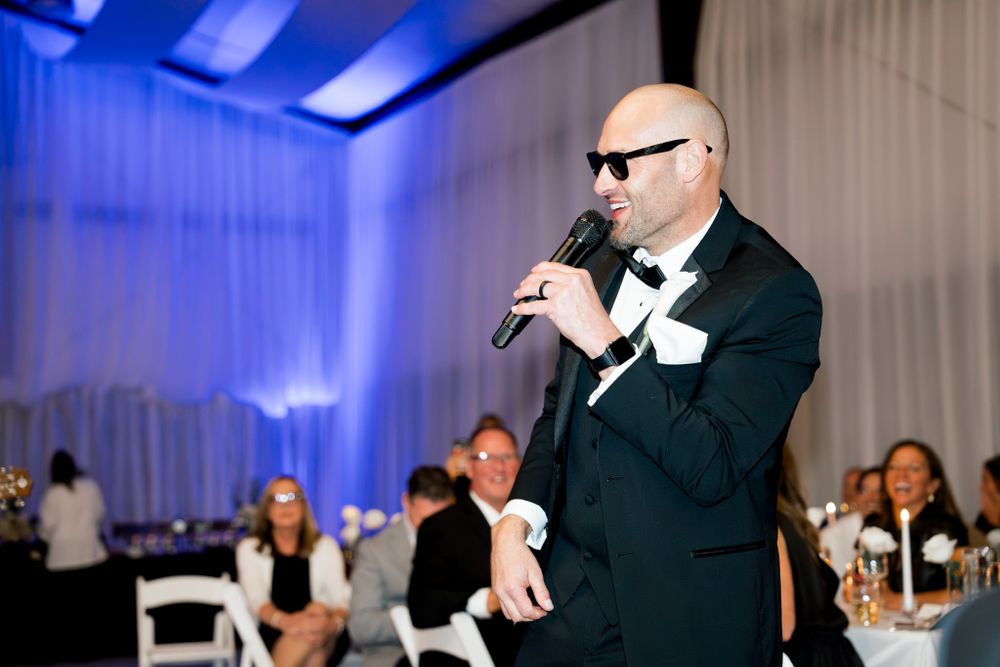 A speaker in a tuxedo addresses guests at an elegant reception with blue lighting.