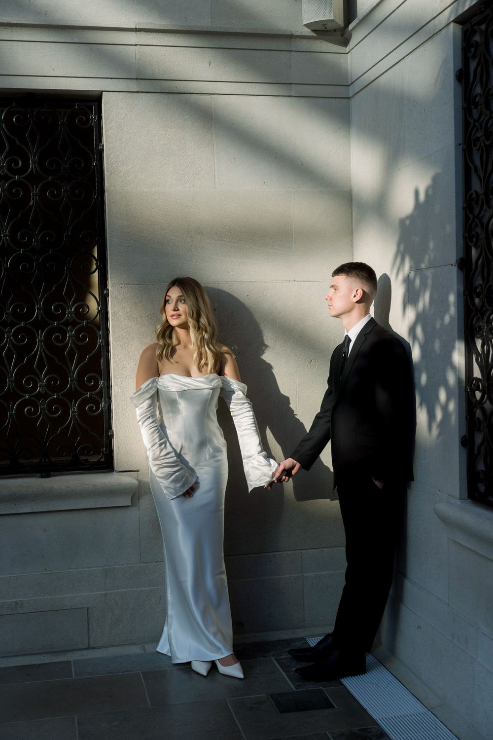 Elegant couple in formal attire pose against a sunlit wall with dramatic shadows.