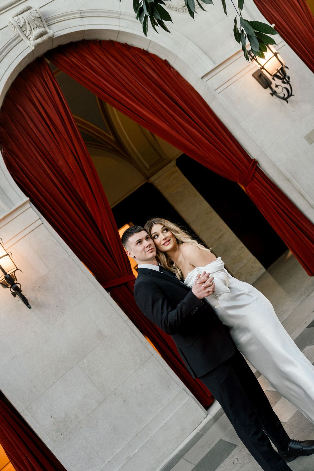 Formal couple poses under an ornate archway with red velvet curtains and classical architecture.