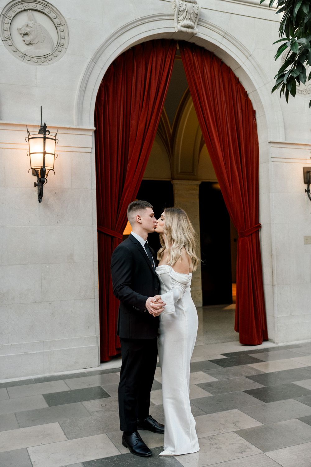 A couple poses romantically beneath an ornate archway with red curtains and wall sconces.