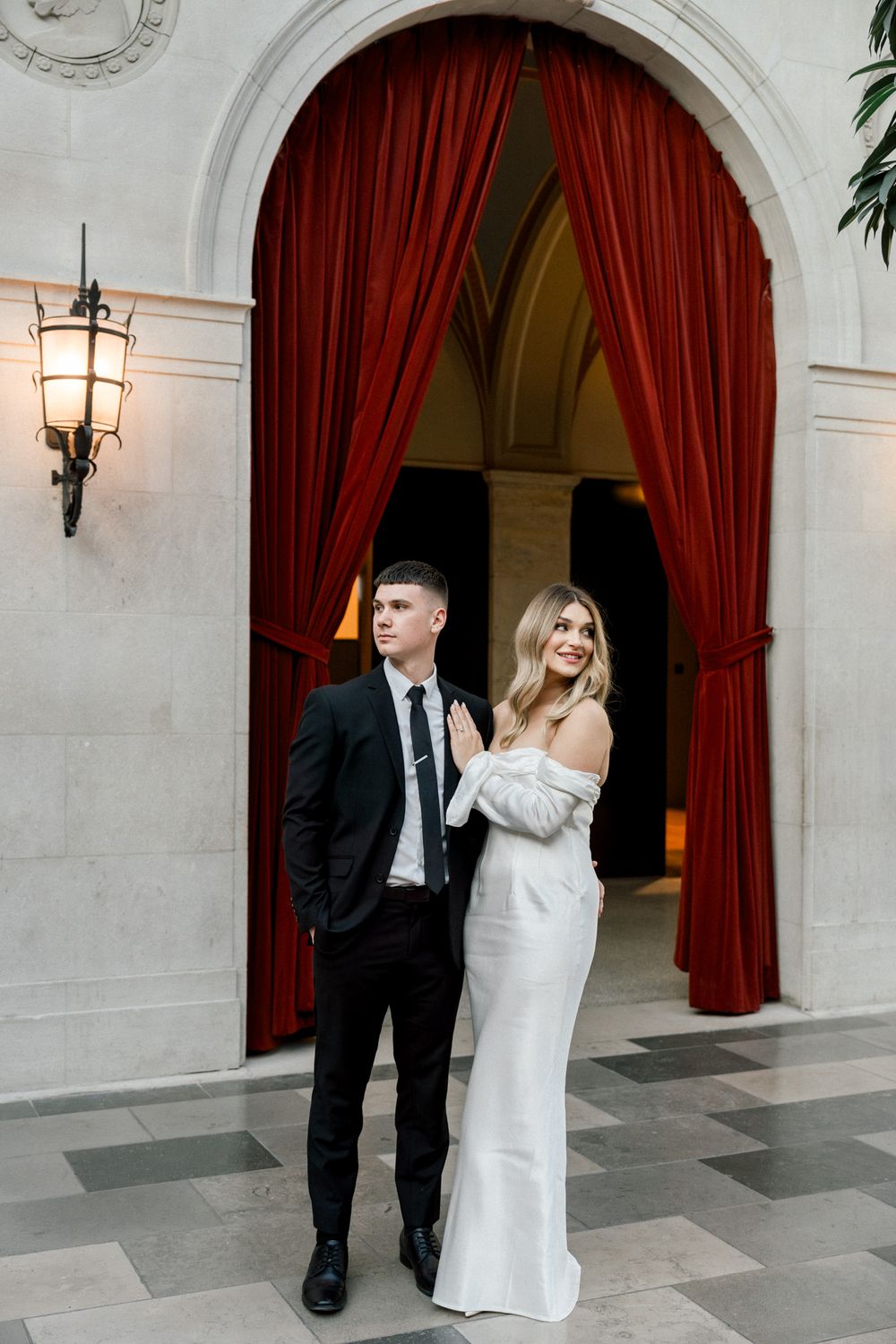 A couple in formal attire poses beneath an elegant archway with dramatic red drapes.