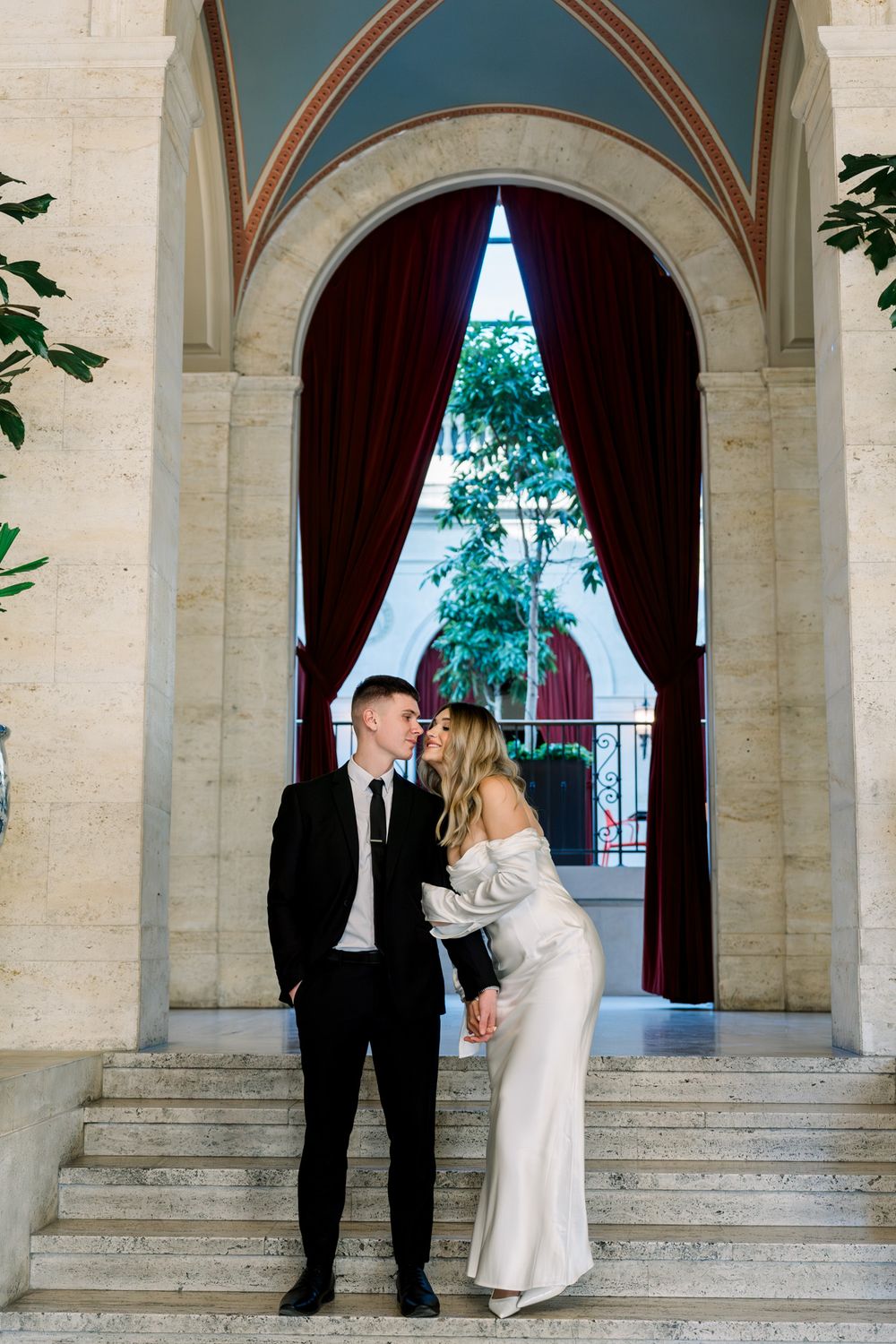 A couple shares an intimate moment on marble steps beneath an arched doorway with red velvet curtains.