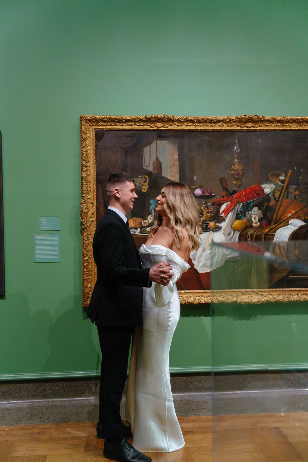 A couple poses in formal attire beside a classical painting with ornate gold frame against green walls.