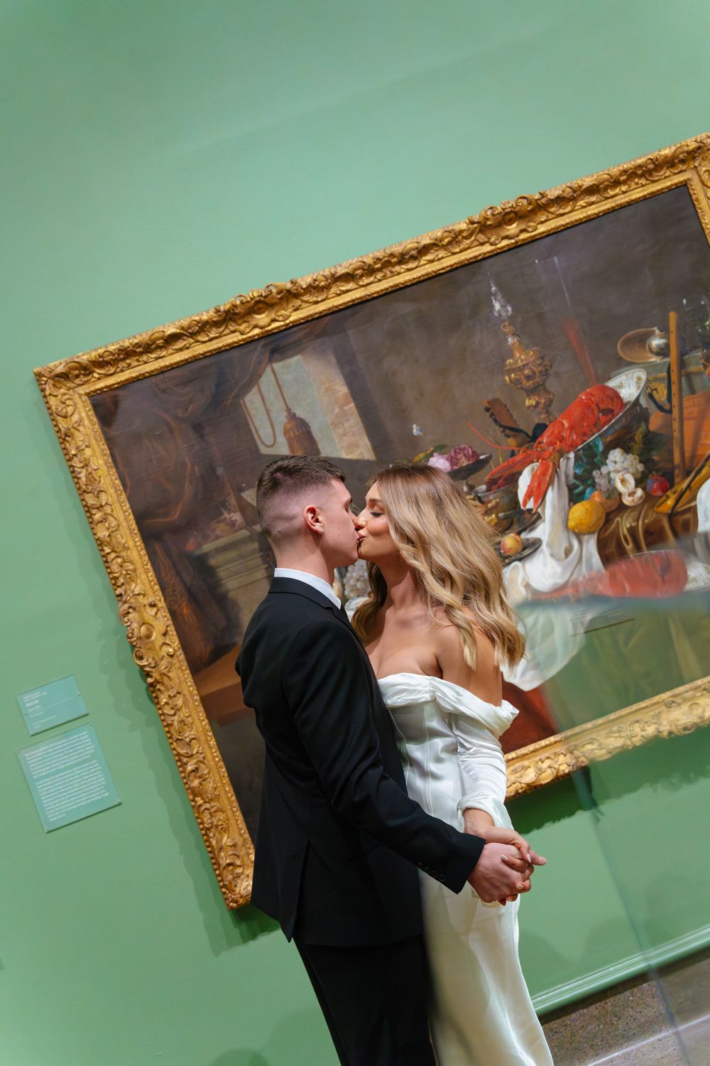 A romantic moment shared in front of an ornate gold-framed painting on a mint green gallery wall.