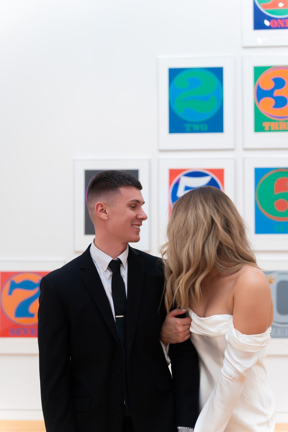 Couple sharing tender moment in art gallery with vibrant numbered prints on wall.